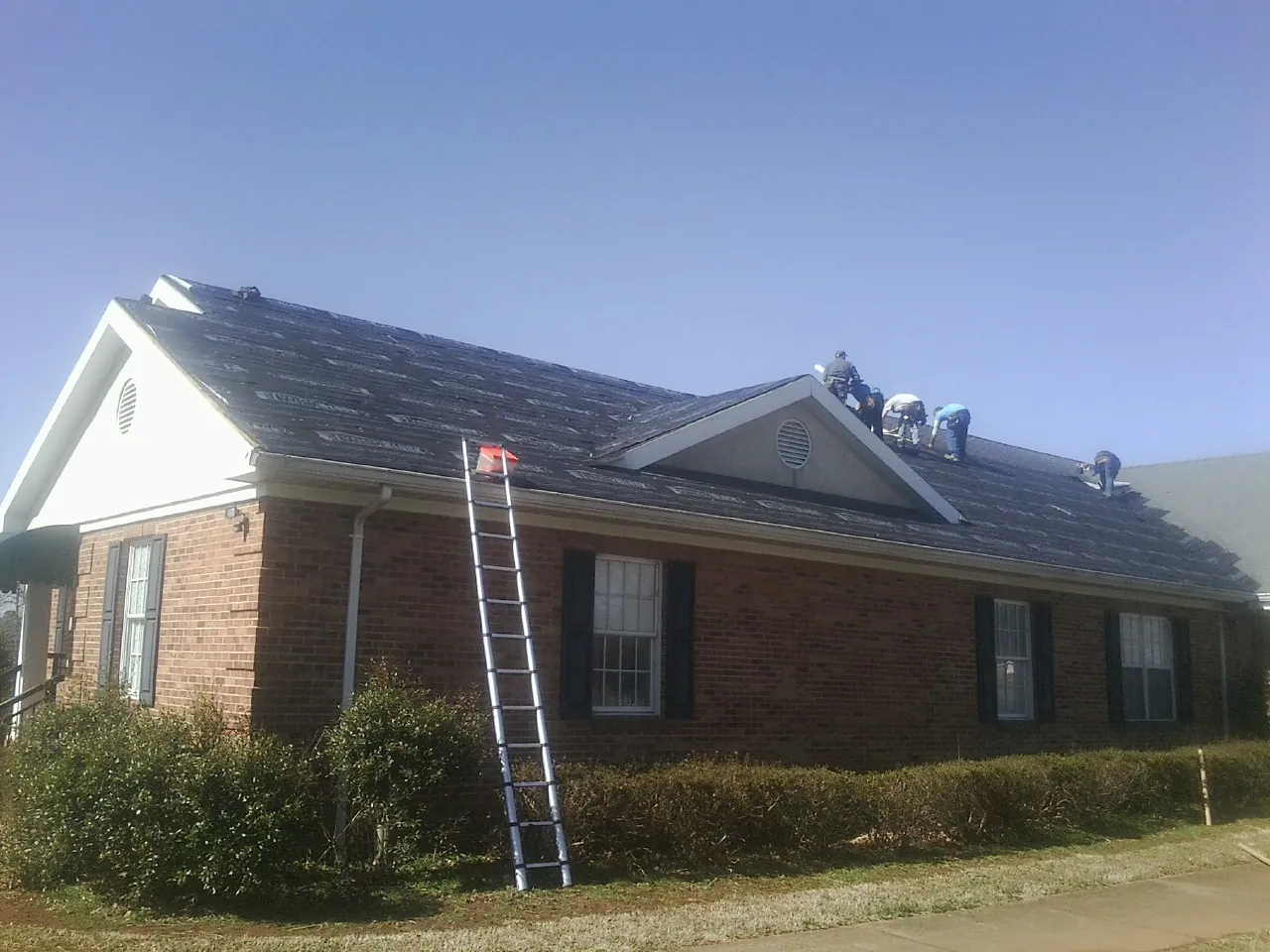 Crew tearing shingles off a brick ranch roof with a ladder set — Emergency Roof Repair work by our crew in Pompey