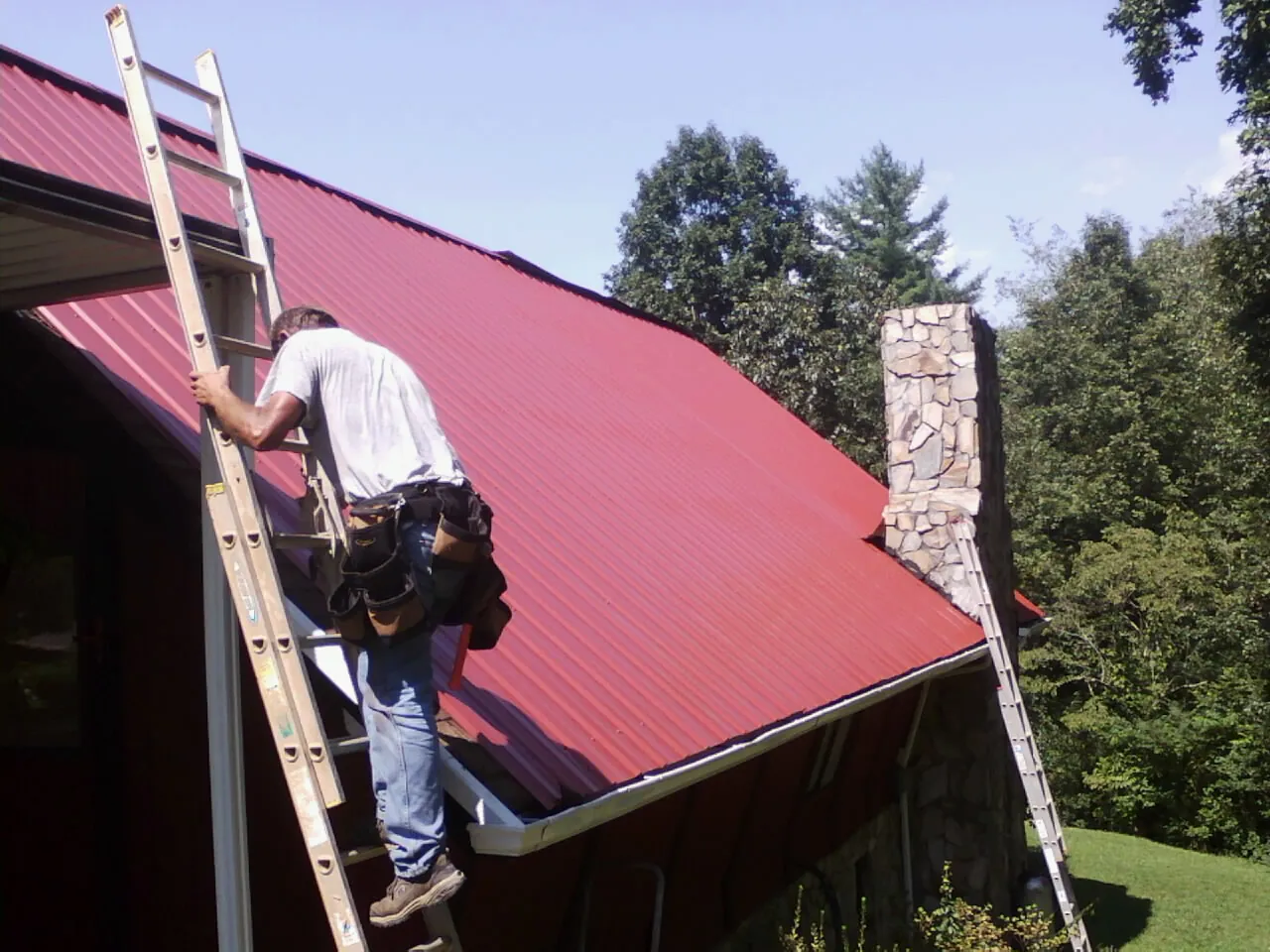 Roofer on a ladder beside a red metal roof and stone chimney — Soffit & Fascia Repair by Roofing Brothers in Pompey